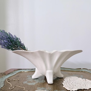 May include: A white ceramic bowl with a scalloped, wavy rim and three short, sturdy feet. The bowl has a smooth, matte finish and contains a sprig of purple lavender. The design features a wide, open top that tapers down to the base.
