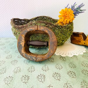 May include: An antique wooden hat stretcher with a dark handle, set against a moss-covered basket with a yellow flower and lavender sprigs. The stretcher is weathered, showing signs of age and use. A wooden shoe last is also visible.
