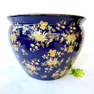 May include: A large, round, navy blue ceramic planter with a gold floral pattern. The planter has a wide opening and a slightly flared rim. The gold floral design is intricate and covers the entire surface of the planter.