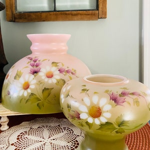 May include: Two hand-painted glass lampshades with a floral design. The larger shade is pink with white and purple flowers. The smaller shade is green with white and purple flowers.