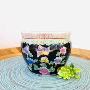 May include: A small black ceramic planter with a floral design featuring pink, yellow, blue, and purple flowers and butterflies. The planter has a white rim with a red and gold design.