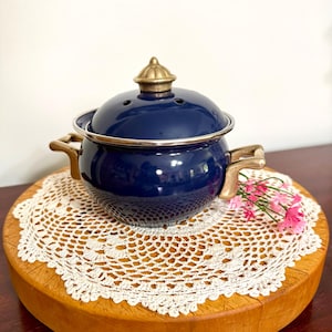 May include: A deep blue enamel pot with a matching lid featuring a metallic gold-toned finial and two side handles. The pot has a silver-toned rim and small ventilation holes on the lid, presenting a rounded, classic kitchenware design.