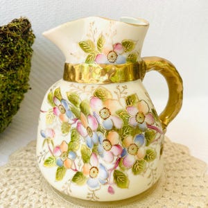 May include: A small, cream-colored ceramic pitcher with a gold handle and trim. The pitcher is adorned with hand-painted floral designs in shades of pink, blue, and orange, with green leaves. The pitcher is a decorative item.