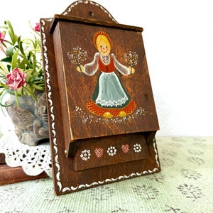 May include: A vintage wooden wall-mounted box with a hand-painted folk art design. The box features a brown finish, a decorative border, and a painted image of a woman in traditional attire. The woman is holding flowers. The box is likely used for storage.