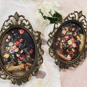 May include: Two oval-shaped ornate gold picture frames with floral designs. The frames are on a white and pink fabric background. The frames contain vintage floral prints.