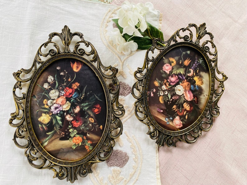 May include: Two oval-shaped ornate gold picture frames with floral designs. The frames are on a white and pink fabric background. The frames contain vintage floral prints.