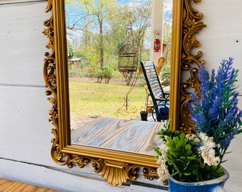 May include: A gold ornate mirror with a detailed, scrolled frame. The mirror reflects a view of a backyard with a birdcage and a chair.