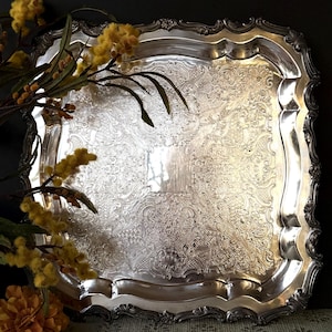 May include: A silver-colored, rectangular serving tray with ornate, embossed floral and scrollwork patterns. The tray has a scalloped edge and a reflective surface. Yellow flowers and greenery are in the foreground, adding a touch of color and contrast.