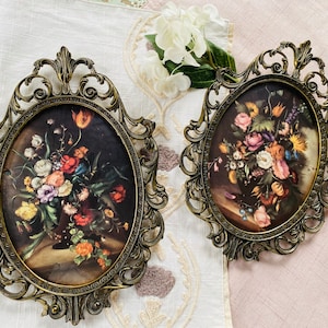 May include: Two ornate gold-toned oval picture frames with intricate scrollwork designs. Each frame holds a vintage floral print with a bouquet of colorful flowers.