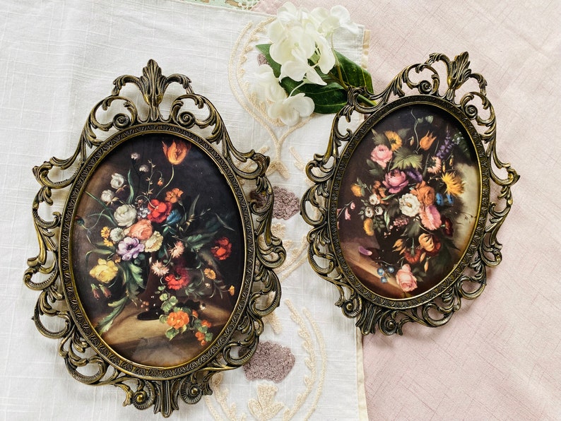 May include: Two ornate gold-toned oval picture frames with intricate scrollwork designs. Each frame holds a vintage floral print with a bouquet of colorful flowers.