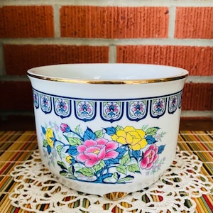 May include: White ceramic planter with a gold rim and a floral design featuring pink, yellow, and blue flowers. The planter has a blue and white geometric pattern around the top.