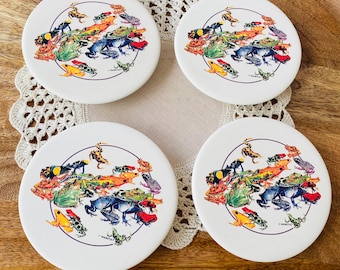 May include: Set of four white ceramic coasters with a colorful design of poison dart frogs. Each coaster features a circle of frogs in various shades of green, blue, yellow, orange, and red.