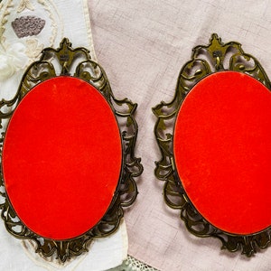 May include: Two ornate gold-colored picture frames with red velvet backing. The frames have a detailed, intricate design.