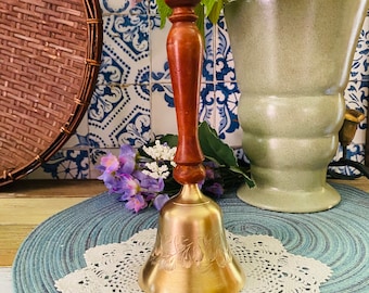 May include: A gold bell with a wooden handle, sitting on a blue and white crocheted doily. The bell has a floral design and is on a blue and white placemat.