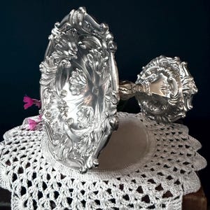 May include: Ornate silver-colored serving dish and goblet with detailed floral and scrollwork designs. The dish is oval-shaped and rests on a white crocheted doily. The goblet is on the right side of the image. The background is a dark blue.