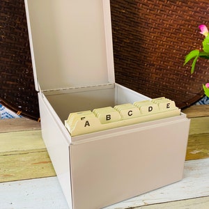 May include: A light gray metal index card box with dividers labeled A through E. The box is open and the dividers are visible.