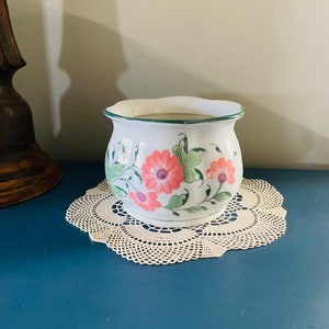 May include: White ceramic planter with a green rim and floral design featuring pink and green flowers. The planter is sitting on a white crocheted doily.