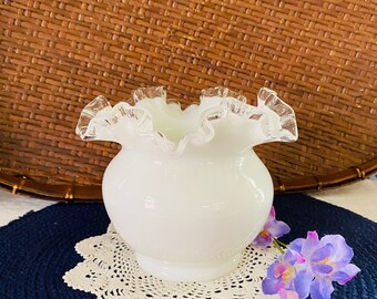 May include: A white milk glass vase with a ruffled edge. The vase is sitting on a white crocheted doily on a blue surface.
