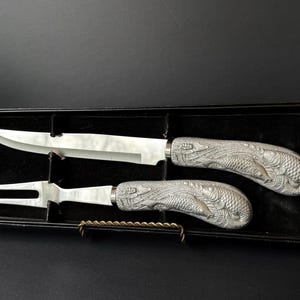 May include: A carving set with a knife and fork, presented in a black box. The silver-toned handles are intricately detailed with a fish scale design. The knife blade is long and slender, and the fork has two tines.