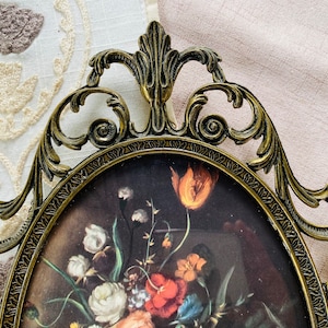 May include: An ornate gold-toned metal picture frame with a detailed floral design. The frame is oval shaped and has a glass front. The frame is empty and ready to display a picture or artwork.