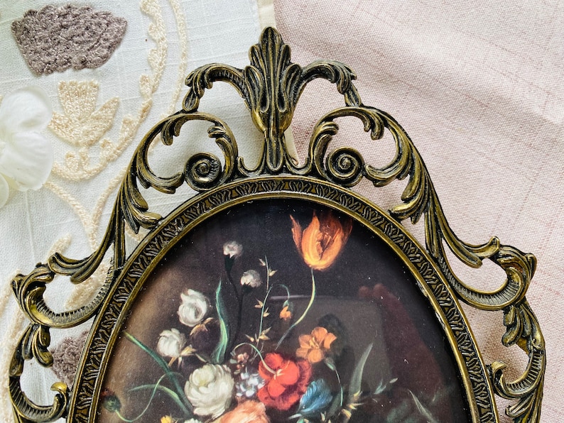 May include: An ornate gold-toned metal picture frame with a detailed floral design. The frame is oval shaped and has a glass front. The frame is empty and ready to display a picture or artwork.