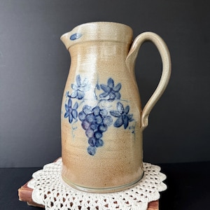 May include: Tan stoneware pitcher with a curved handle and spout. The pitcher is decorated with blue grape and floral designs. It sits on a white lace doily and a stack of books. The pitcher has a rustic, vintage aesthetic.
