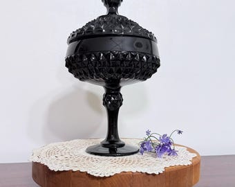 May include: A black glass compote dish with a lid, featuring a raised diamond pattern on the bowl and lid, a tall pedestal base with a matching diamond-textured accent, and a pointed finial top, standing as a decorative glass vessel.