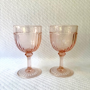 May include: Two pink-tinted, stemmed glasses. The glasses have a rounded bowl with vertical ridges and a decorative pattern. The stem is slender, and the base is a circular, ridged design. The glasses are set against a white background.