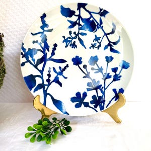 May include: White ceramic plate with a blue floral design. The plate features a delicate watercolor-style pattern of blue flowers and foliage. The plate is displayed on a gold-colored stand, with a small green plant in front.