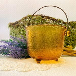 May include: A textured, amber-colored glass bucket with a dark metal handle. The bucket has three small feet and a decorative, textured surface. The background includes lavender sprigs and a mossy decorative element.