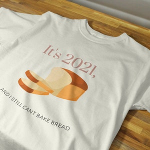 May include: Cream-coloured t-shirt with the text "It's 2021, and I still can't bake bread". The shirt has a graphic of a loaf of bread with two slices in pink and brown tones.