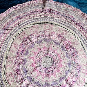 Könnte beinhalten: Rund gestrickter Schal in Weiß-, Lavendel- und Rosatönen. Der Schal weist komplizierte Muster und Texturen auf, mit einem zentralen Blumenmotiv und konzentrischen Kreisen, die sich nach außen ausdehnen. Die sanfte Farbpalette schafft ein zartes und elegantes Accessoire.
