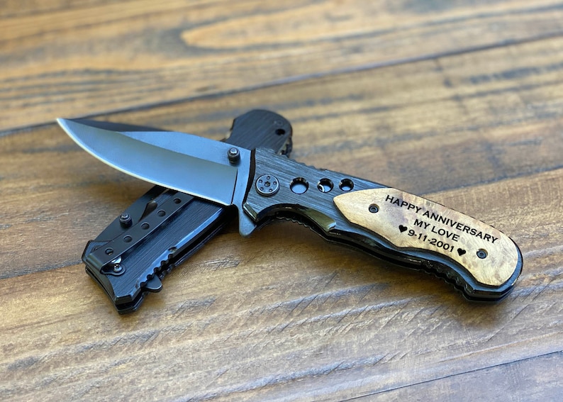May include: A black folding pocket knife with a wood handle. The handle is engraved with the text "Happy Anniversary My Love 9-11-2001" and two hearts.