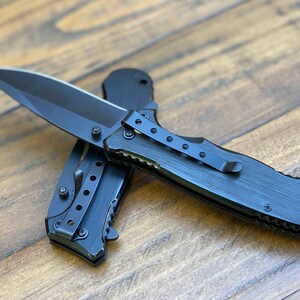 May include: A black folding knife with a black handle and a partially serrated blade. The knife has a pocket clip and a textured handle.
