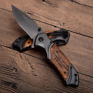 May include: A folding pocket knife with a dark gray blade and a wooden handle. The handle has the engraved text "YOU'LL ALWAYS BE MY GREATEST CATCH! 5.20.2021". The knife is set against a wooden background.