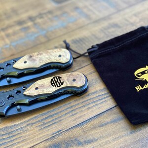 May include: Two black pocket knives with wood handles and a monogrammed "ABC" on the handle. The knives are in a black velvet pouch with a gold embroidered logo that says "Blackknife" and a cartoon ninja.