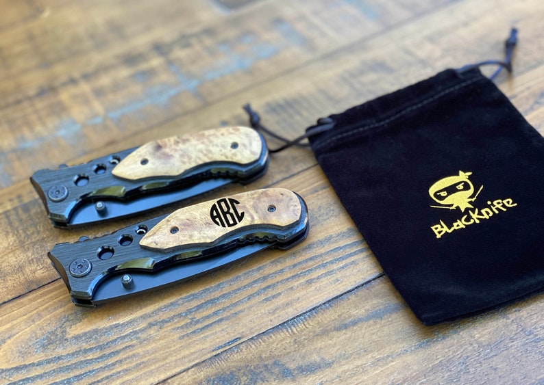 May include: Two black pocket knives with wood handles and a monogrammed "ABC" on the handle. The knives are in a black velvet pouch with a gold embroidered logo that says "Blackknife" and a cartoon ninja.