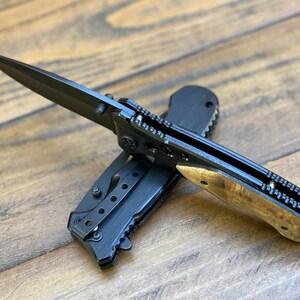 May include: A black folding pocket knife with a wood handle. The blade is open and the knife is lying on a wooden surface.