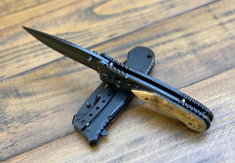 May include: A black folding pocket knife with a wood handle. The blade is open and the knife is lying on a wooden surface.