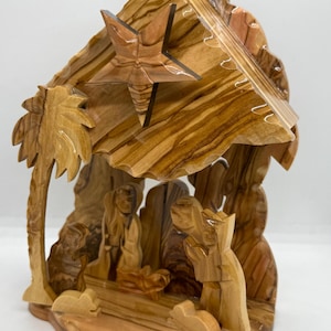 Puede incluir: Un belén de madera tallada que representa la Sagrada Familia en un establo. La escena está hecha de madera de color marrón claro y presenta una estrella, una palmera y figuras de María, José y el niño Jesús. El diseño general es detallado y artístico.