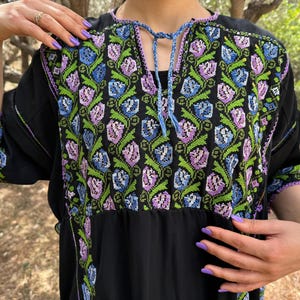 May include: Black dress with floral embroidery in shades of blue, green, and purple. The embroidery is concentrated on the bodice and sleeves, with a drawstring neckline. The dress has short sleeves and a flowing skirt.