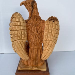 Op de afbeelding: Een gebeeldhouwde houten adelaarssculptuur met uitgespreide vleugels. De adelaar is gedetailleerd met getextureerde veren en staat op een rechthoekige houten basis. De sculptuur heeft tinten licht- en donkerbruin.