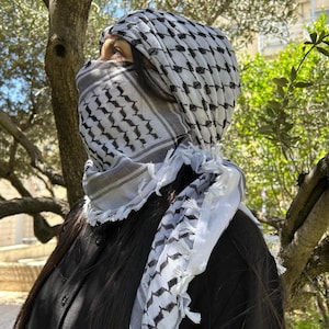 May include: A black and white keffiyeh scarf, draped over the head and shoulders. The scarf features a geometric pattern and fringed edges. The person is wearing a black shirt, with long dark hair. The background includes green foliage and a tree.