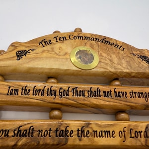 May include: A wooden plaque inscribed with "The Ten Commandments" in black lettering. The plaque is made of light-coloured wood and includes a small, round, gold-coloured inset. The first two commandments are also engraved.