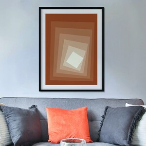 May include: A gray sofa with pillows in various colors and textures, including a white pillow with a fringe, a black pillow, and an orange pillow. A framed abstract print with brown and beige squares hangs on the wall above the sofa. A glass vase with a green plant sits on a coffee table in front of the sofa.