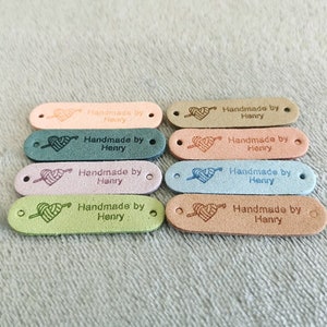Pode incluir: Dez etiquetas de couro ovais com um design de cora&ccedil;&atilde;o e fio. Cada etiqueta diz "Handmade by Henry". As etiquetas s&atilde;o de v&aacute;rias cores, incluindo rosa, verde, azul e marrom.