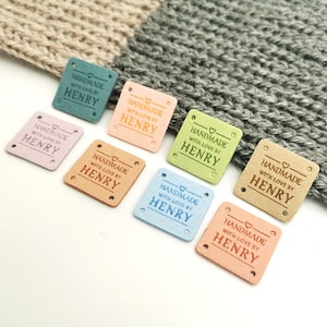 May include: A selection of small, square tags in pastel shades, each bearing the inscription "HANDMADE WITH LOVE BY HENRY". The tags, in colours like blue, pink, and green, have two holes for attaching to handmade items.