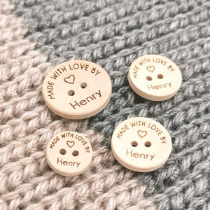 May include: Three wooden buttons with the text "Made with love by Henry" and a small heart symbol. The buttons are different sizes and are on a knitted fabric background.