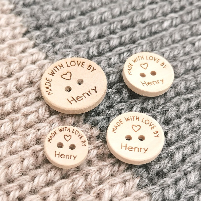 May include: Three wooden buttons with the text "Made with love by Henry" and a small heart symbol. The buttons are different sizes and are on a knitted fabric background.