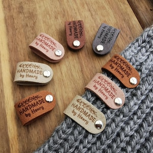 May include: Assortment of eight leather cord keepers in various shades of brown, beige, and grey. Each keeper features a silver metal button and is engraved with the text "HANDMADE by Henry" in a brown font.